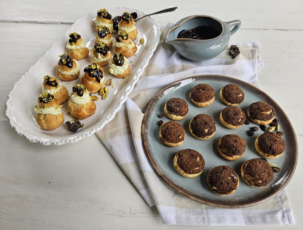 A platter of cream-filled pastries topped with chocolate and pistachios, alongside a round plate of coffee-flavored cookies, with a small jug of chocolate sauce in the background.