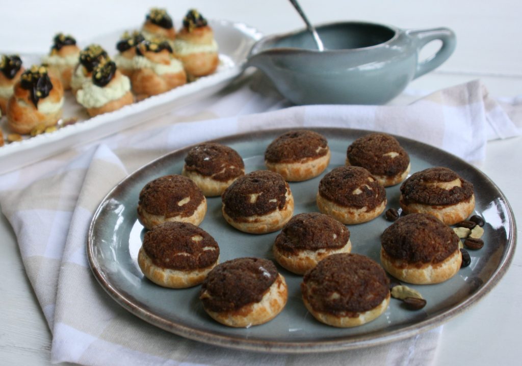 A close-up view of a plate filled with small pastry items topped with a chocolate or coffee-like topping, surrounded by additional pastries in the background. A small bowl with a sauce and a white napkin are also visible.