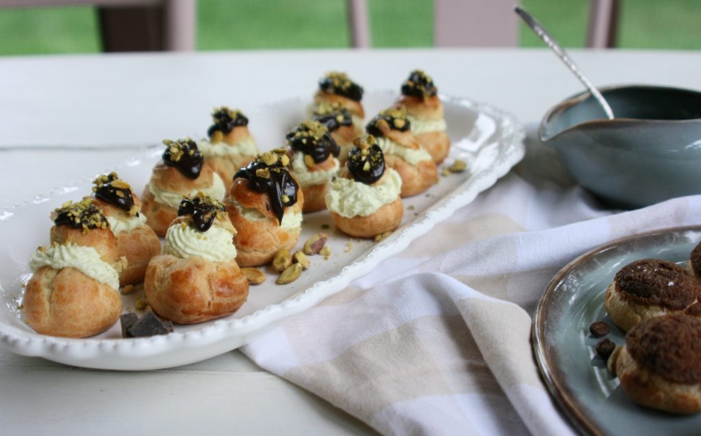A platter of cream-filled pastry puffs topped with chocolate and crushed pistachios, with a small dish of sauce nearby.