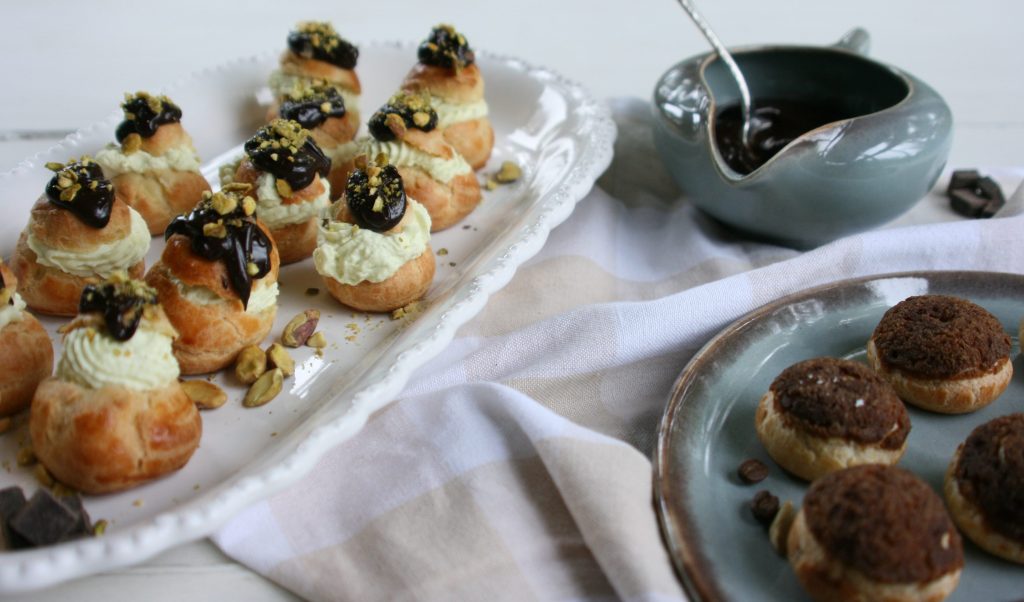 A platter of filled cream puffs topped with chocolate ganache and crushed nuts, accompanied by a bowl of chocolate sauce and a plate of round chocolate-filled pastries.